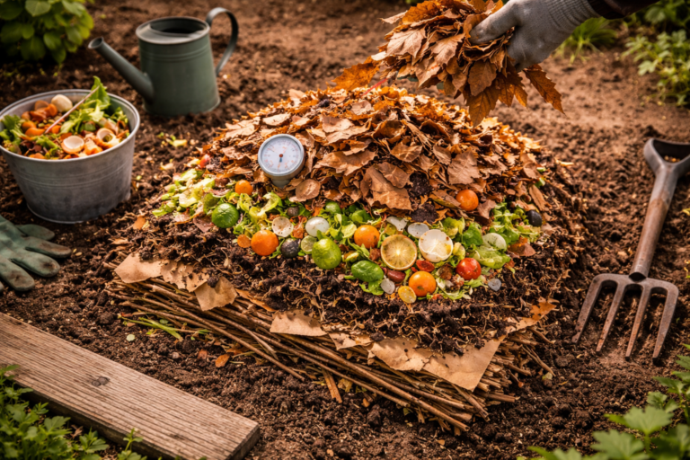 Compost pile being built showing layers of sticks, kitchen scraps, and dry leaves with a compost thermometer, garden fork, and watering can